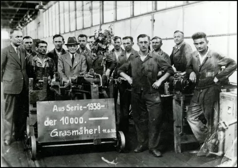 Schwarz weiß foto von Mitarbeitern von früher mit dem Schild: Aus Serie 1938 - der 10.000 Grasmäher