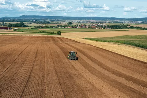 Fendt 500 Vario fährt mit Anbaugerät über weites Feld mit Dörfern im Hintergrund