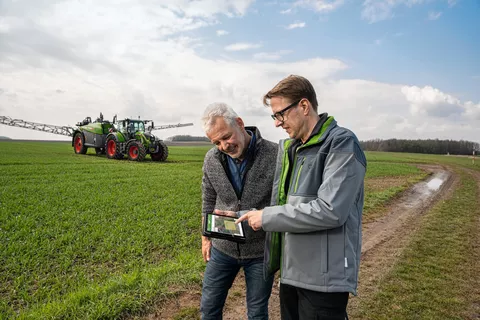 Zwei Landwirte prüfen bei FendtONE auf dem IPad die Daten des hinter Ihnen stehenden Fendt Rogator 300