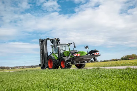 Fendt 314 Vario fährt bei schönem Wetter mit Heck- und Frontmähwerk auf einer Straße neben einer Wiese