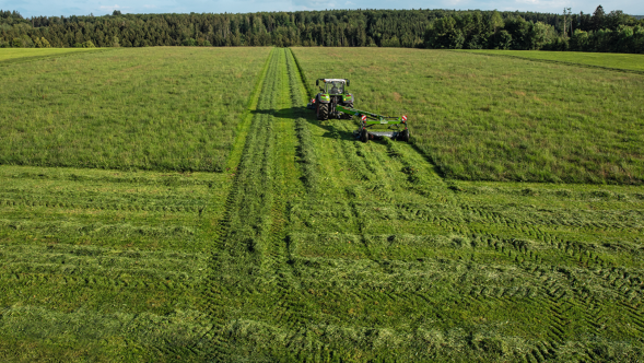 Fendt Slicer gezogen mit Transportfahrwerk sowie Frontmähwerk im Grünland. Im Hintergrund ist Wald zu sehen.