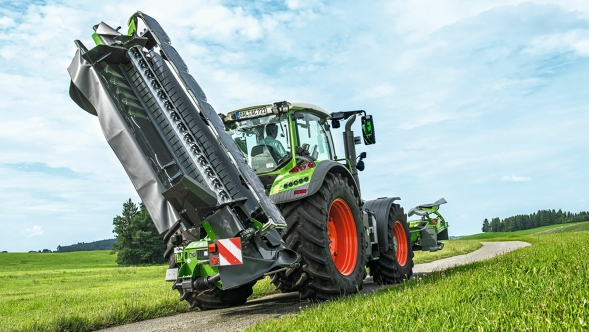 Ein grüner Fendt Vario Traktor mit Fendt Slicer TLX in Transportstellung auf dem Feldweg. Umher sind grüne Wiesen und blauer Himmel mit Wolken bedeckt.