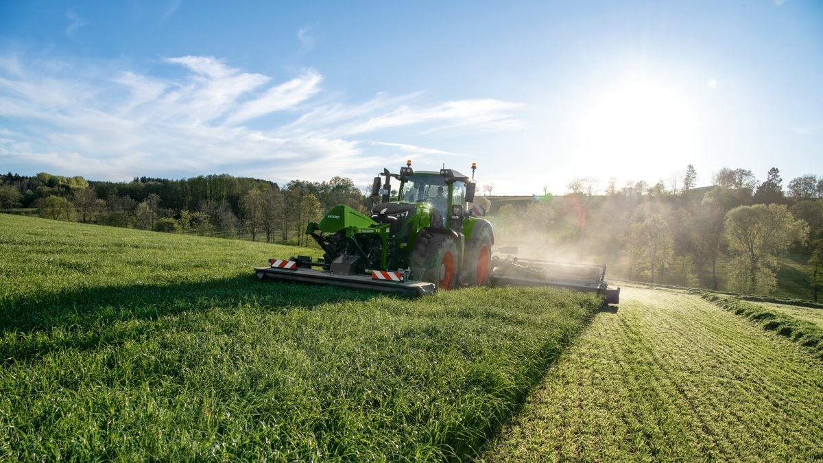 Ein Landwirt mäht mit einem Fendt Slicer Front- und Heckmähwerk ein grünes Feld bei bewölktem Himmel.