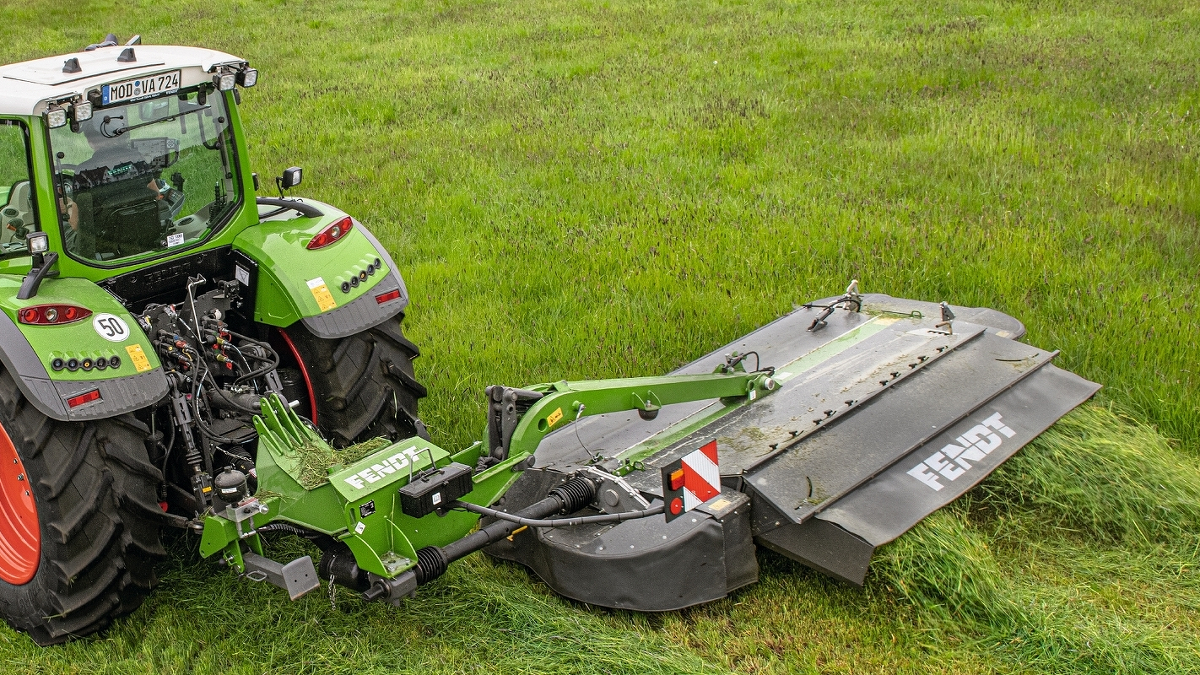 Ein Landwirt mäht mit einem Fendt Slicer Front- und Heckmähwerk ein grünes Feld bei bewölktem Himmel.