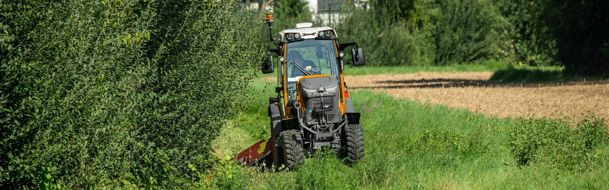 Ein orange lackierter ISU Fendt e100 V Vario im Kommunaleinsatz an einem Grünstreifen