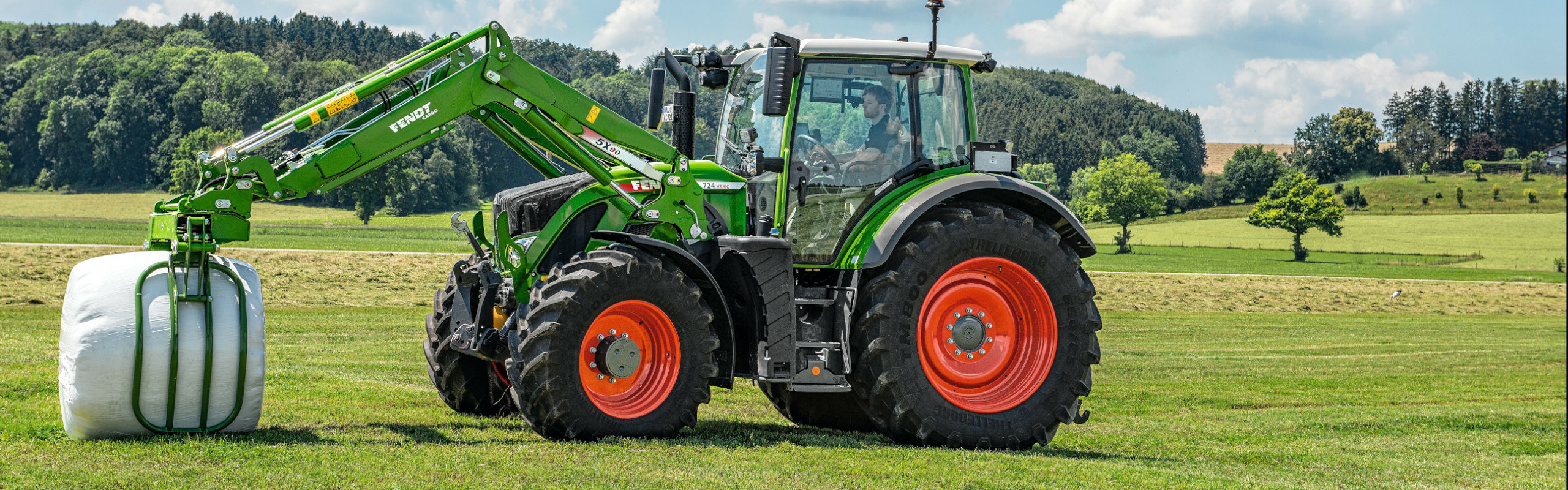 Ein Landwirt steht mit einem Fendt 700 Vario Gen6 auf einer grünen Wiese und greift mit dem Fendt Cargo Frontlader einen weißen Siloballen.