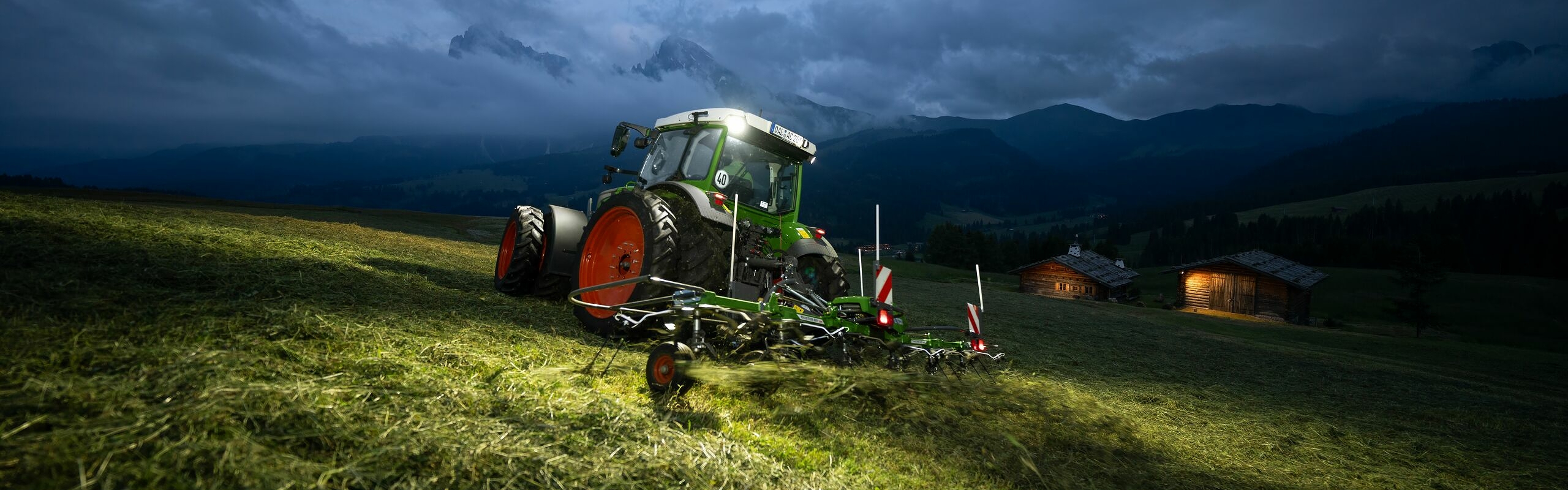 Der grüne Fendt Traktor mit roten Felgen und dem grünen Anbaugerät Fendt Twister fährt in Schräglage auf einer Wiese in den Bergen. Im Hintergrund sind Wolken, Berge und eine beleuchtete Hütte zu sehen.