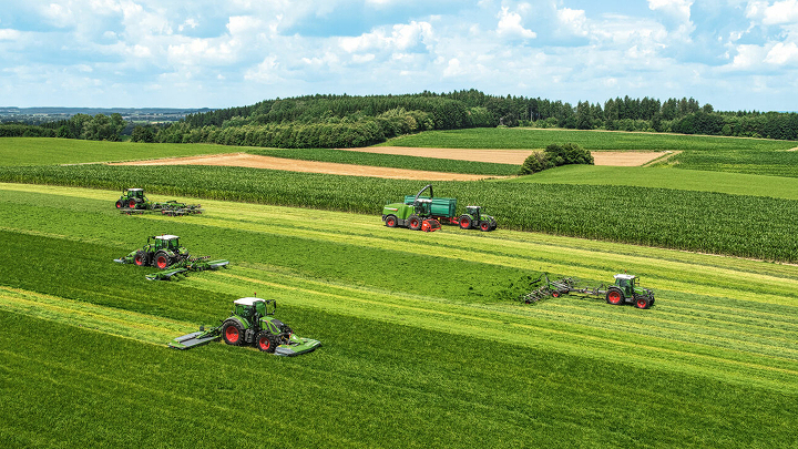 Six Fendt tractors and a Fendt Katana forage harvester driving in a staggered formation across a green field, mowing, turning, swathing and chopping.