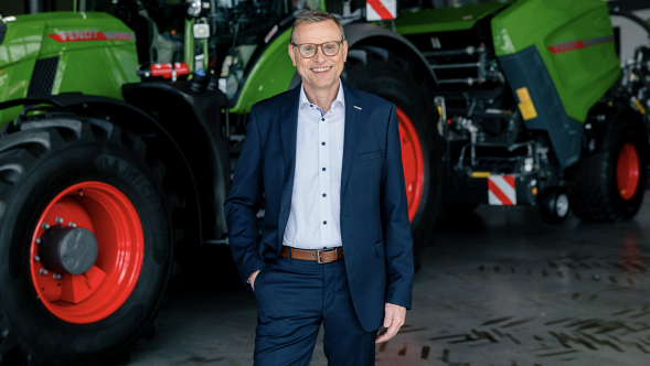 Walter Wegner, de terno azul escuro, sorri em frente a um trator Fendt com uma enfardadeira de fardos redondos.