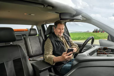 Landwirt sitzt im Auto auf sein Handy schauend mit Fendt Traktor im Hintergrund