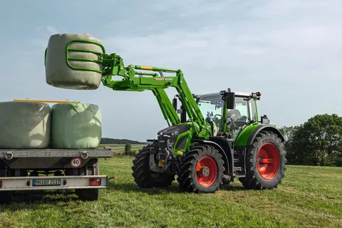 Ein Fendt Traktor mit einer Ballengabel, lädt einen eingepackten Heuballen auf einen Anhänger