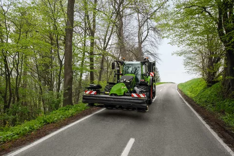 Ein Fendt Traktor mit einem Fendt Mähwerk fährt auf einer Landstraße durch den Wald.