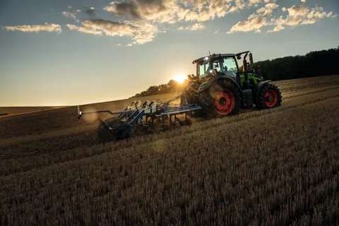 Ein Fendt Traktor mit einem Grubber fährt bei Sonnenuntergang über ein Stoppelfeld