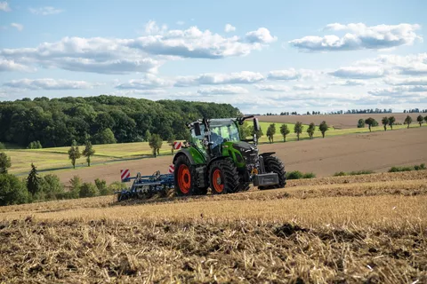 Ein Fendt Traktor mit einem Grubber auf einem Feld mit blauem Himmel und Wolken im Hintergrund.