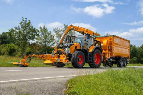 Ein von vorne rechts fotografierter Fendt 728 Vario in Kommunal-orange mäht mit einem Auslegemäher den Straßenrand und zieht einen Anhänger