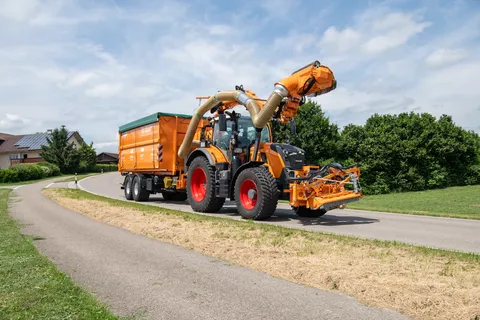 Ein kommunal-oranger Fendt 728 Vario fährt mit einem Auslegermäher und einem Anhänger auf einer Straße im Transport, im Hintergrund eine Ortschaft