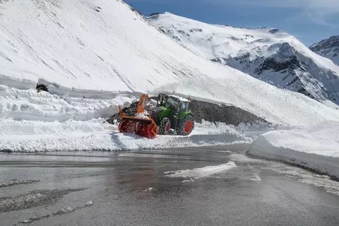 Ein Fendt 728 Vario in grün fährt mit Schneeketten und einer Schneefräse durch hohen Schnee um die Straße freizufräsen im Hintergrund verschneite Berge