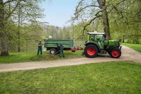 Zwei Mitarbeitende in der Landschaftspflege beladen einen Anhänger hinter einem Fendt e107 Vario mit Blättern in einem Park mit einem von Bäumen umringten See