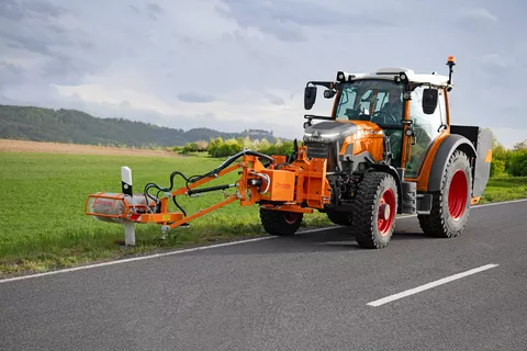 Ein Fendt e107 Vario in kommunal-orange mit einem Frontanbaugerät zur Reinigung von Leitpfosten fährt auf der Straße und reinigt Leitpfosten