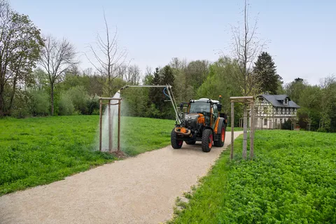 Ein Fendt e107 Vario in kommunal-orange bewässert mit einem an der Front des Traktors angebauten Gießarm einen Baum in einem Park im Frühjahr, im Hintergrund ist ein altes, renoviertes Fachwerkhaus zu sehen