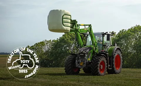 Fendt 620 Vario mit angehobenem Fendt Frontlader auf einer Wieser mit einem Rundballen in der Ballenzange - vorne links ist das Logo desractors of the Year in der Kategorie MidPower in weiß platziert