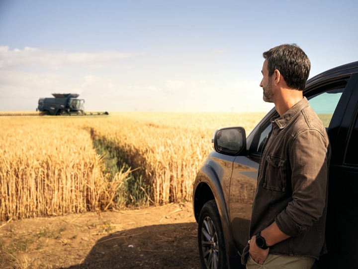 Ein Landwirt steht angelehnt an einem Auto vor seinem Getreidefeld und schaut zufrieden zu, wie ein Fendt Mähdrescher erntet.
