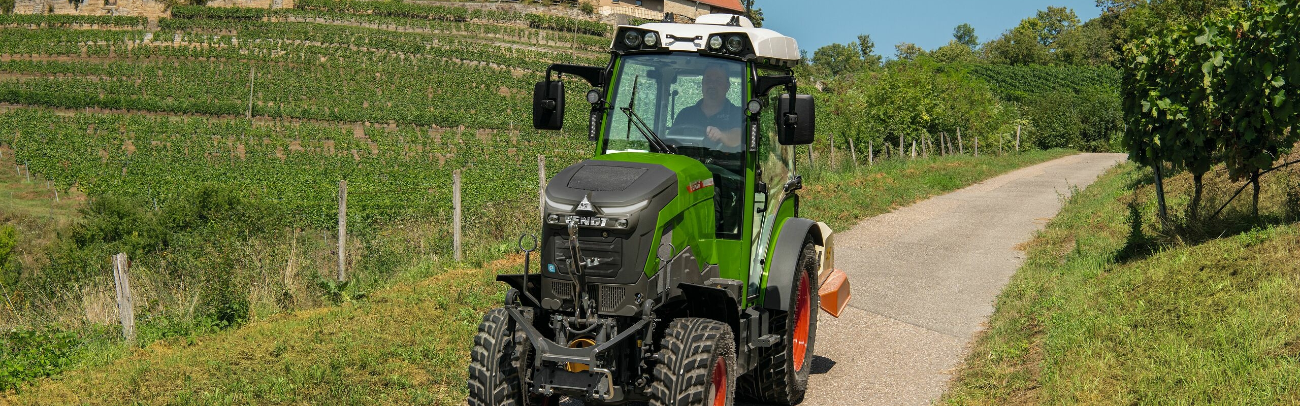 Ein grün lackierter Fendt e100 V Vario fährt zwischen Rebstöcken auf einer schmalen Straße