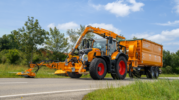 Ein von vorne rechts fotografierter Fendt 728 Vario in Kommunal-orange mäht mit einem Auslegemäher den Straßenrand und zieht einen Anhänger