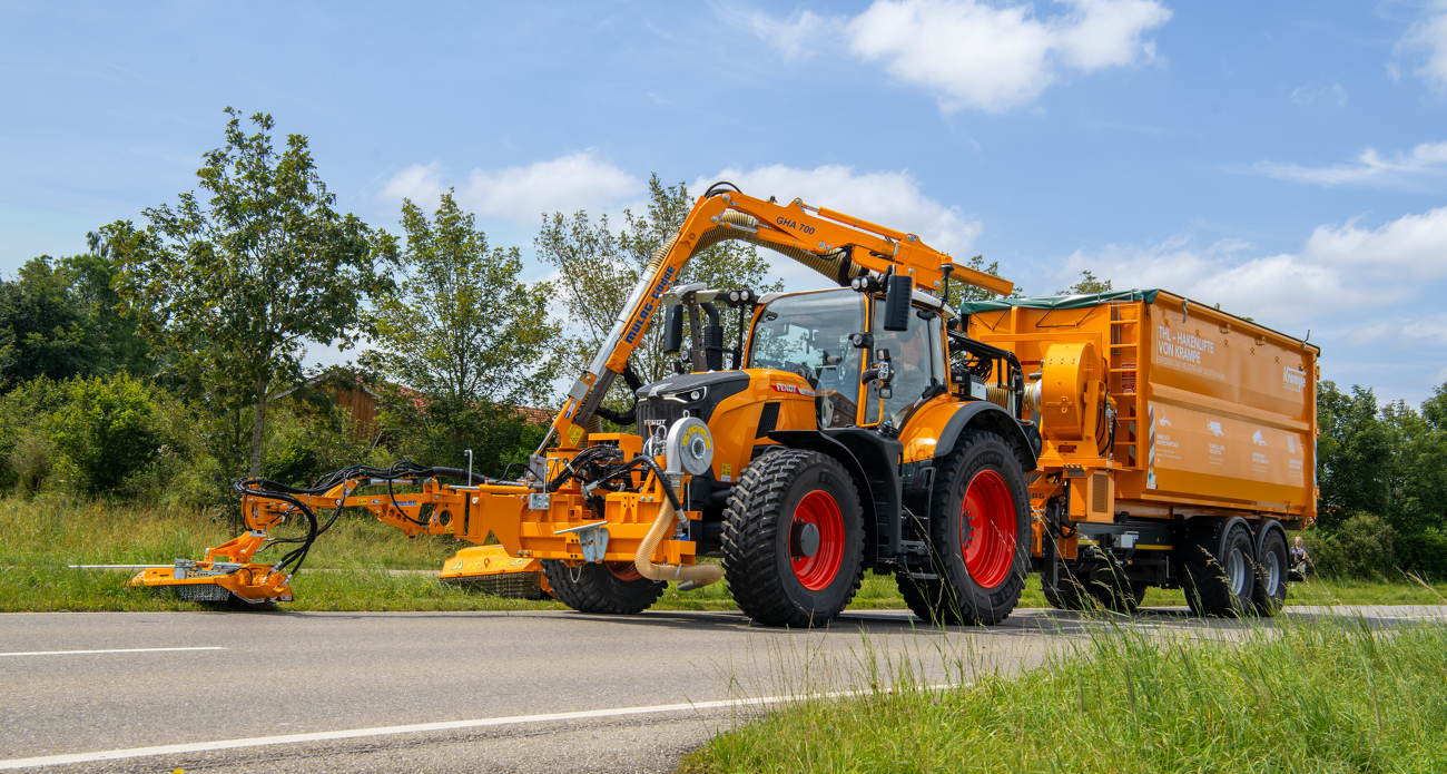 Ein von vorne rechts fotografierter Fendt 728 Vario in Kommunal-orange mäht mit einem Auslegemäher den Straßenrand und zieht einen Anhänger