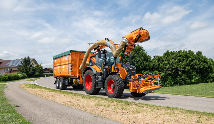 Ein kommunal-oranger Fendt 728 Vario fährt mit einem Auslegermäher und einem Anhänger auf einer Straße im Transport, im Hintergrund eine Ortschaft