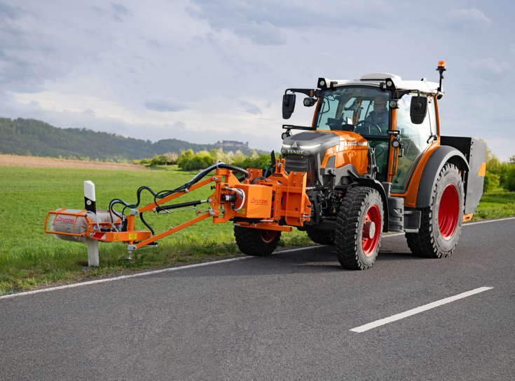 Ein Fendt e107 Vario in kommunal-orange mit einem Frontanbaugerät zur Reinigung von Leitpfosten fährt auf der Straße und reinigt Leitpfosten