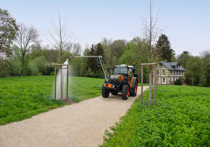 Ein Fendt e107 Vario in kommunal-orange bewässert mit einem an der Front des Traktors angebauten Gießarm einen Baum in einem Park im Frühjahr, im Hintergrund ist ein altes, renoviertes Fachwerkhaus zu sehen