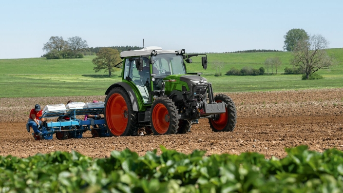 Ein Fendt e100 Vario fährt für Pflanzarbeiten mit einer Pflanzmaschine und Arbeitern im Heck über ein Feld im Vordergrund Erdbeerpflanzen