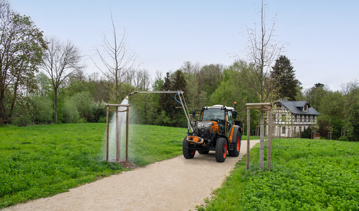 Ein Fendt e107 Vario in kommunal-orange bewässert mit einem an der Front des Traktors angebauten Gießarm einen Baum in einem Park im Frühjahr, im Hintergrund ist ein altes, renoviertes Fachwerkhaus zu sehen