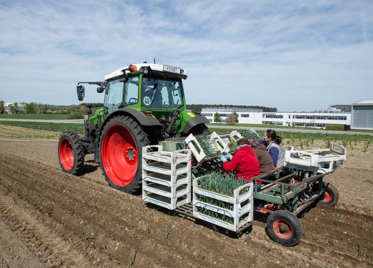 Ein Fendt e107 Vario mit einer Pflanzmaschine für Gemüse beladen mit Gemüsekisten und Pflanzerinnen hinten auf der Maschine