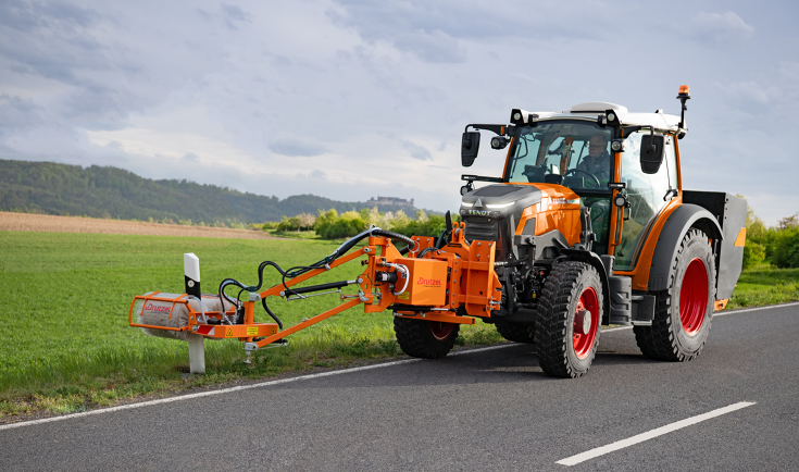 Ein Fendt e107 Vario in kommunal-orange mit einem Frontanbaugerät zur Reinigung von Leitpfosten fährt auf der Straße und reinigt Leitpfosten