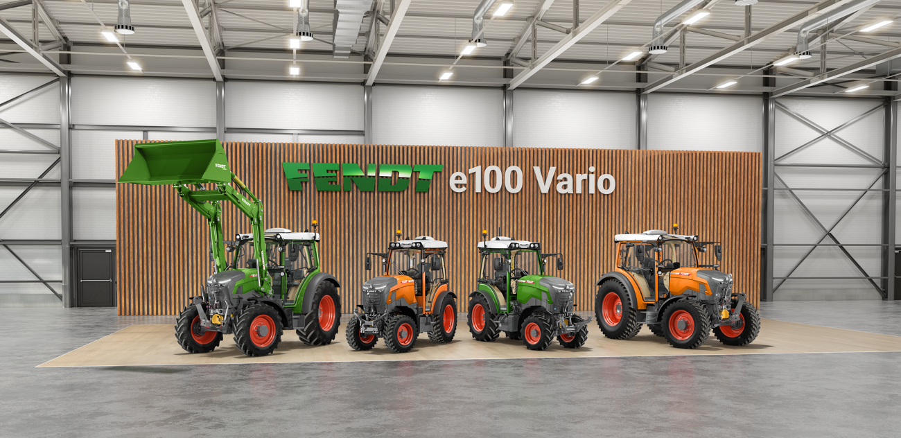 In einer Reihe vor einer Wand mit Holzlatten stehen 4 Modelle des Fendt e100 Vario, v.l. ein grüner Fendt e107 Vario mit Frontlader, ein Fendt e107 V Vario in kommunal-orange, daneben einer in grün, als Vierter ein Fendt e107 Vario in kommunal-orange