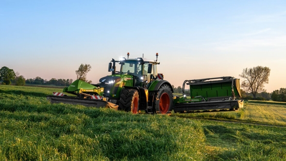 Ein Landwirt fährt bei leichter Dämmerung mit einem Fendt Slicer Front- und Heckmähwerk auf einer grünen Wiese.