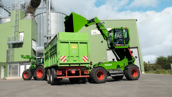 Ein Fendt Teleskoplader mit ausgefahrener Kabine und ausgeklappter Schaufel steht rechts neben einem grünen Anhänger vor einer grünen Lagerhalle und einem silbernen Silo.