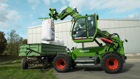 Der ausgefahrene Fendt Cargo T955 belädt vor einer Lagerhalle einen Ladewagen.