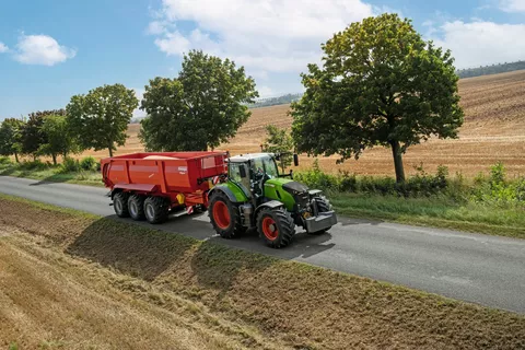 En grøn Fendt 728 Vario med en fuldt lastet Krampe-vogn kører ind på en vej med træer på venstre side