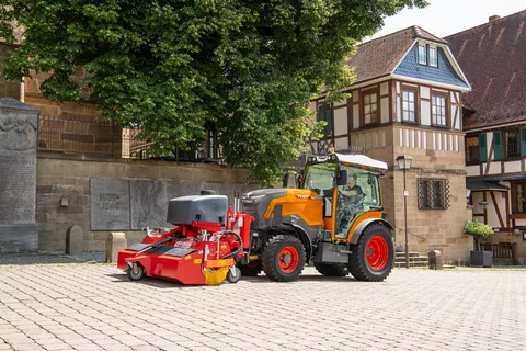 En kommunal orangefarvet Fendt e107 V Vario med en fejemaskine foran fejer fortovet i en by, i baggrunden sandstensmure, et træ og et bindingsværkshus