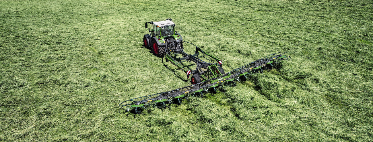 En grøn Fendt Traktor med røde fælge og det grønne Fendt Lotus redskab i arbejde med høst af græs på en grøn eng
