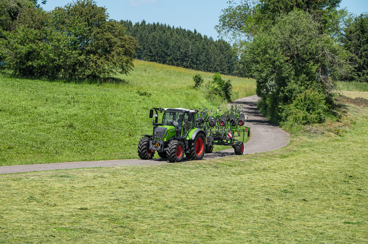 En grøn Fendt Traktor med røde fælge og det grønne Fendt Lotus redskab kører på en smal vej med en grøn eng, blå himmel og udsigt over landskabet.