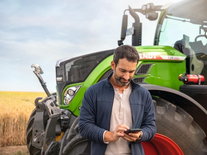 En landmand står ved siden af en Fendt 724 traktor og ser på sin mobiltelefon.