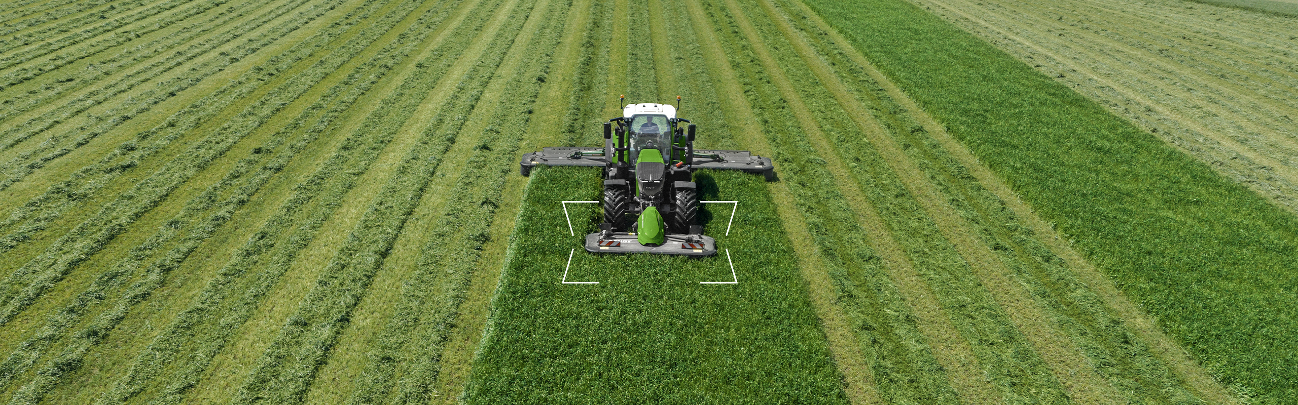 En grøn Fendt 500 Vario med front- og bagmonteret slåmaskine og tændt lys på en eng i morgengryet.