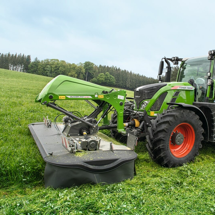 En landmand slår en grøn eng med en Fendt Slicer FQ frontklipper og en Fendt 724 Vario, med træer i baggrunden.