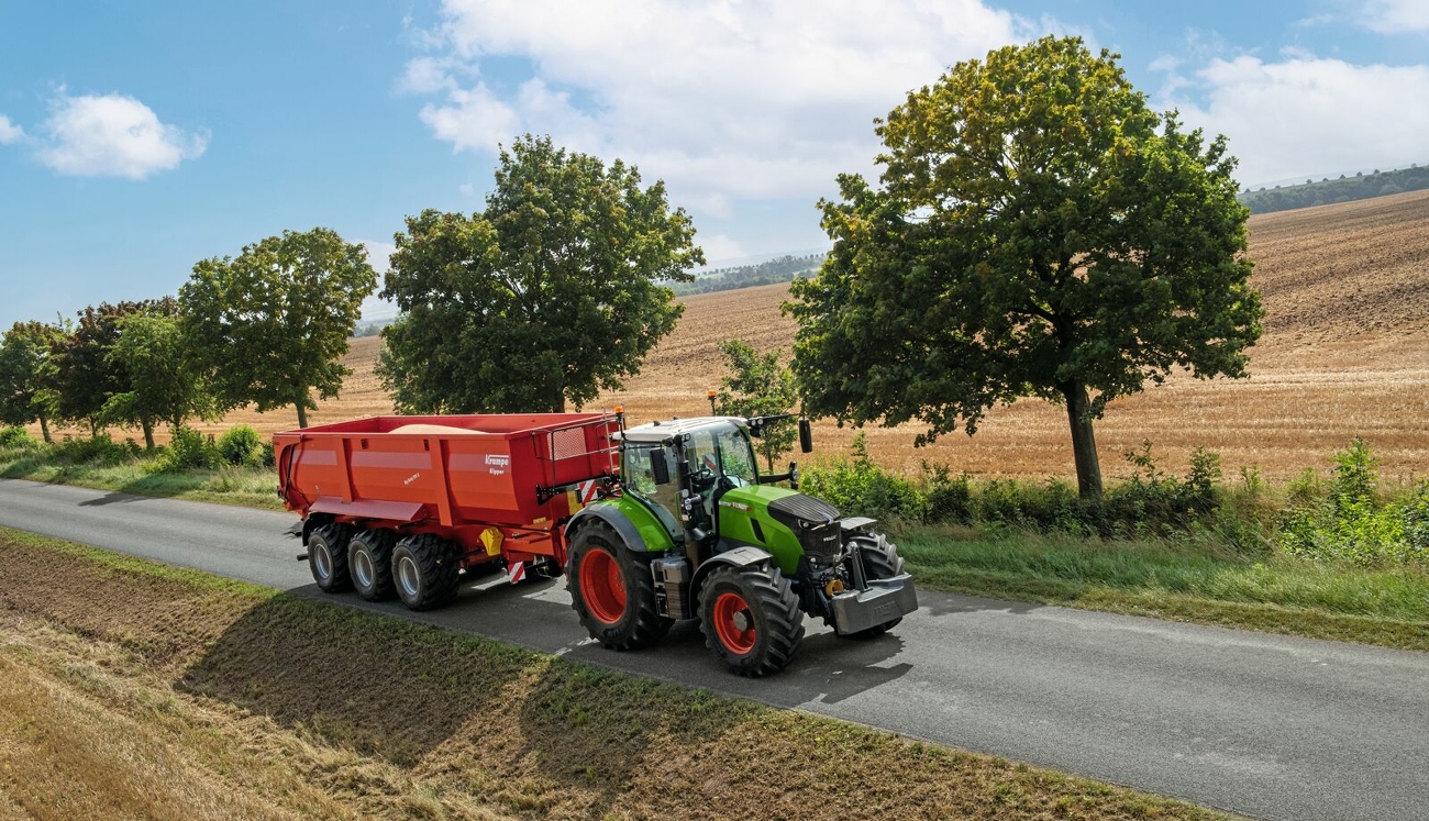 En grøn Fendt 728 Vario med en fuldt lastet Krampe-vogn kører ind på en vej med træer på venstre side