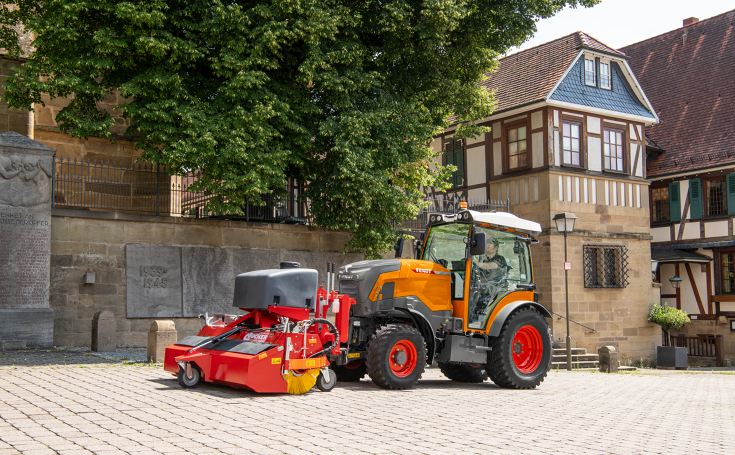 En kommunal orangefarvet Fendt e107 V Vario med en fejemaskine foran fejer fortovet i en by, i baggrunden sandstensmure, et træ og et bindingsværkshus