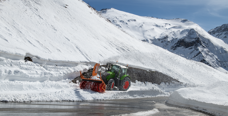 En grøn Fendt 728 Vario kører med snekæder og en sneslynge gennem tung sne for at rydde vejen, med snedækkede bjerge i baggrunden.